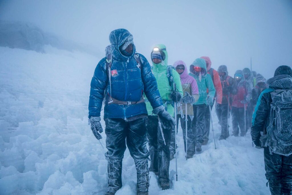 A group of climbers wearing colorful winter gear and headlamps trek through thick snow and fog, covered in frost, on a snowy mountain.