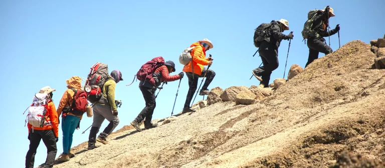 Six hikers wearing backpacks and outdoor gear use trekking poles as they ascend a steep, rocky slope under a clear blue sky.