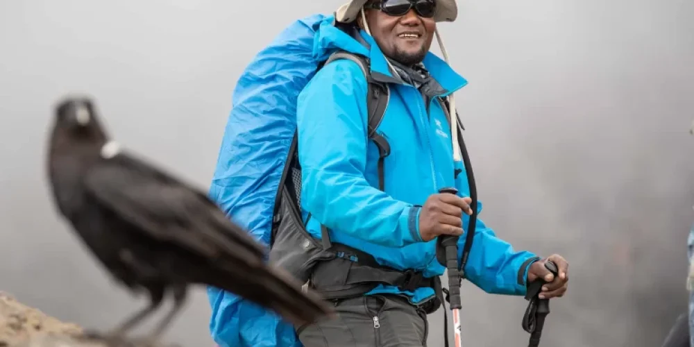 A man in hiking gear and sunglasses smiles while holding trekking poles on a rocky trail. A large black bird is perched on a nearby rock. Misty weather creates a foggy background.